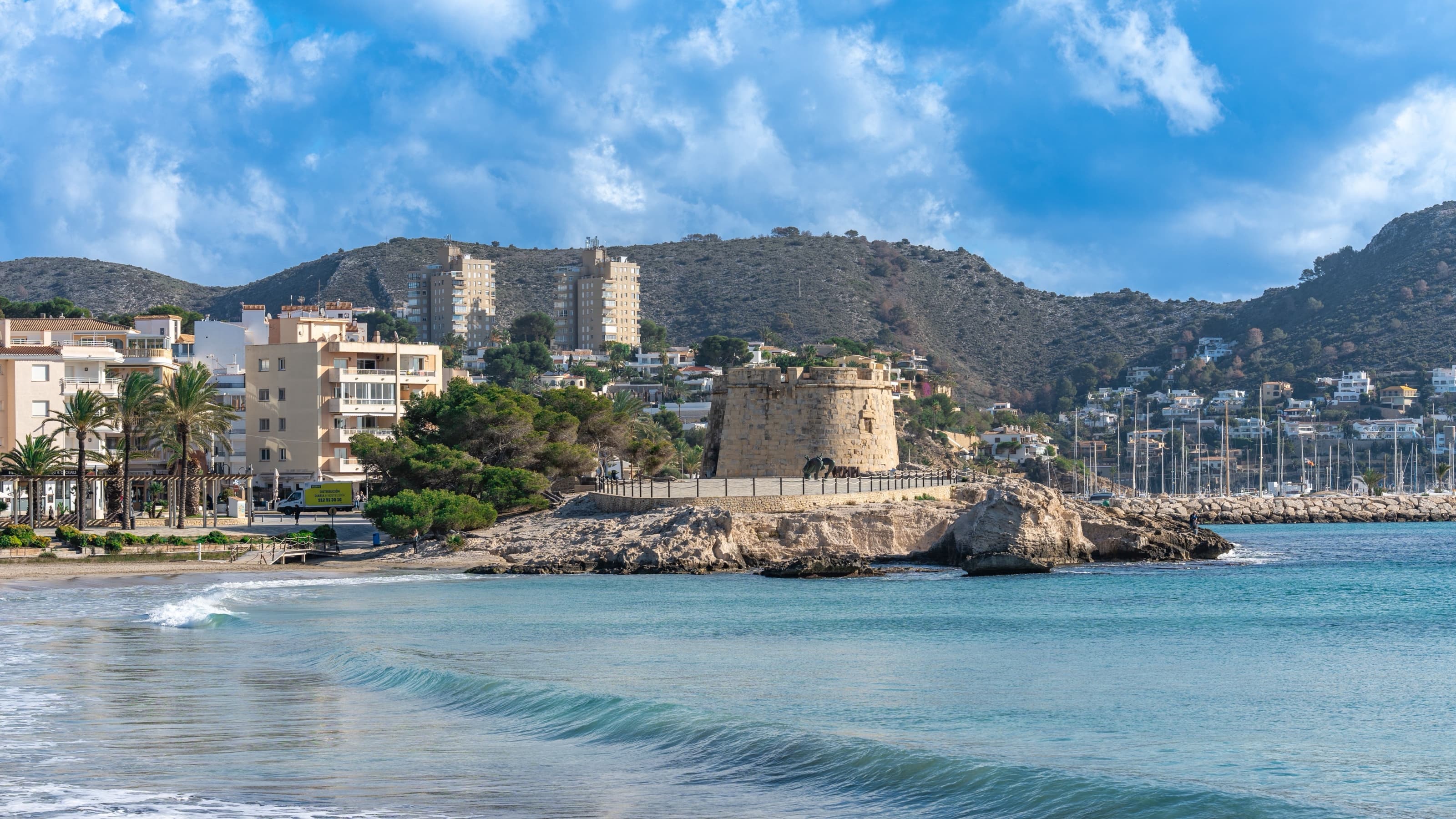 La playa de l'Ampolla de Moraira con el castillo circular del siglo XVIII y los mástiles del puerto deportivo detrás